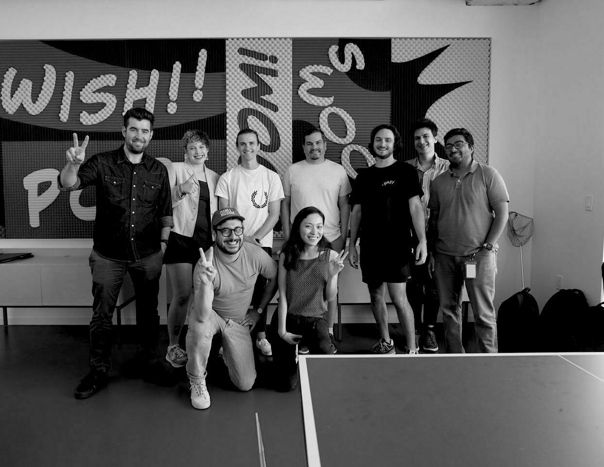 Auto-generated description: A group of ten people are posing together, some gesturing with peace signs, in front of a wall with graphic text.