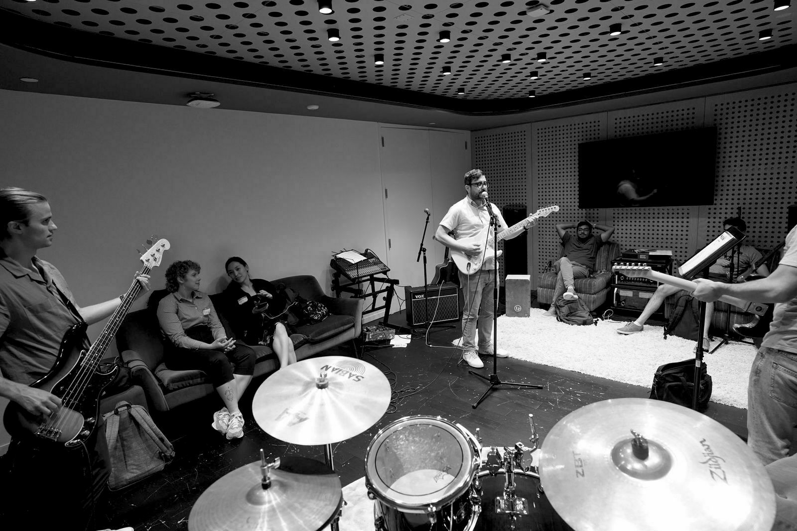 Auto-generated description: A group of musicians is rehearsing in a studio, with drums in the foreground and people sitting and playing instruments.